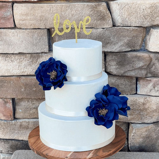 Three-tiered white cake with blue flowers and a 'love' topper against a stone wall.