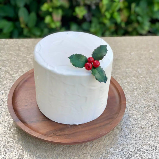Holly leaves made from gumpaste sitting on top of a white one tier cake. The cake is on a round wooden cake stand.