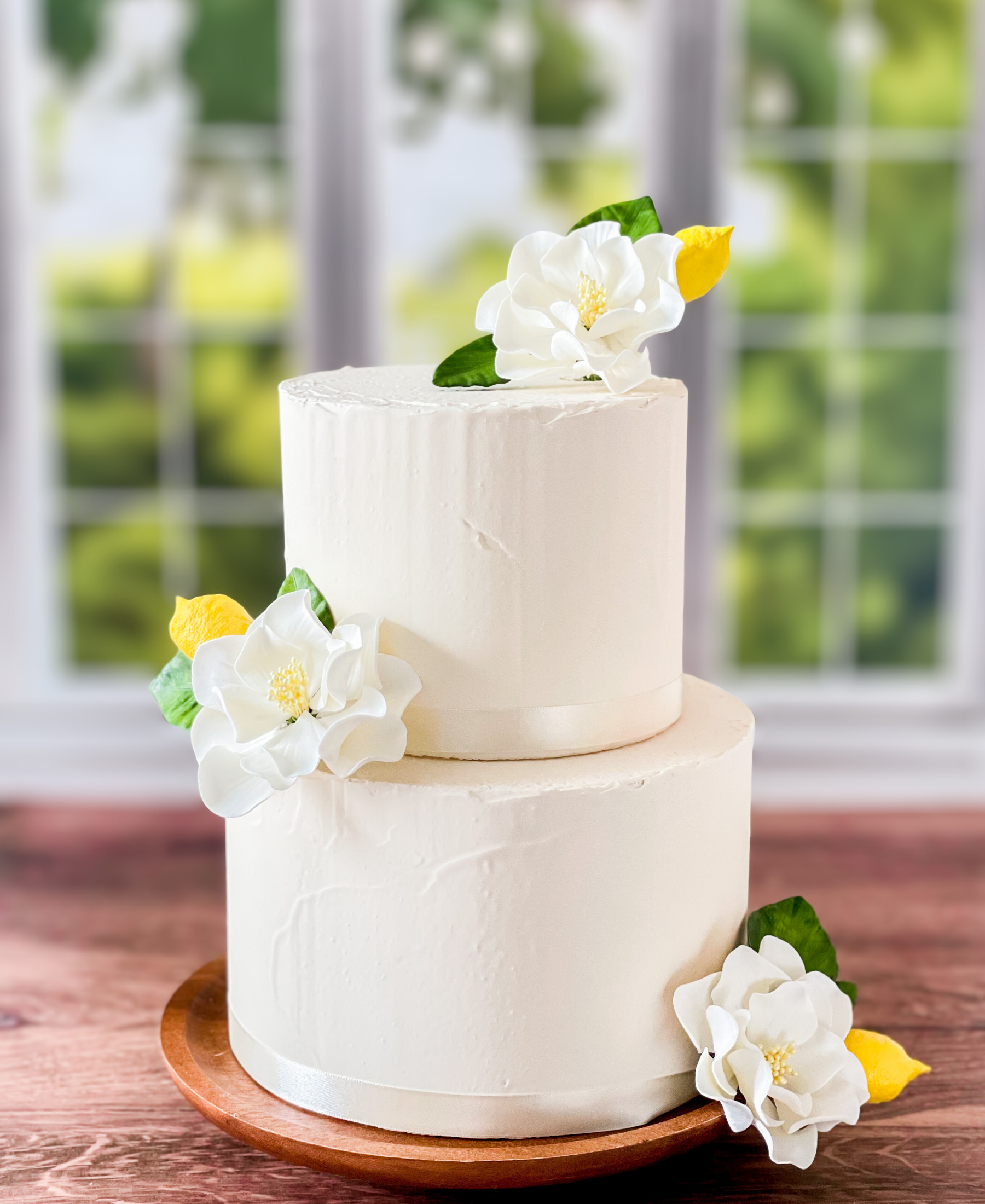 Two-tiered white cake with sugar floral and Gumpaste lemon decorations on a wooden stand.