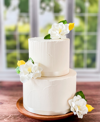Two-tiered white cake with sugar floral and Gumpaste lemon decorations on a wooden stand.