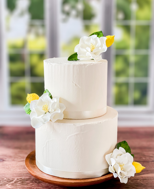 Two-tiered white cake with sugar floral and Gumpaste lemon decorations on a wooden stand.
