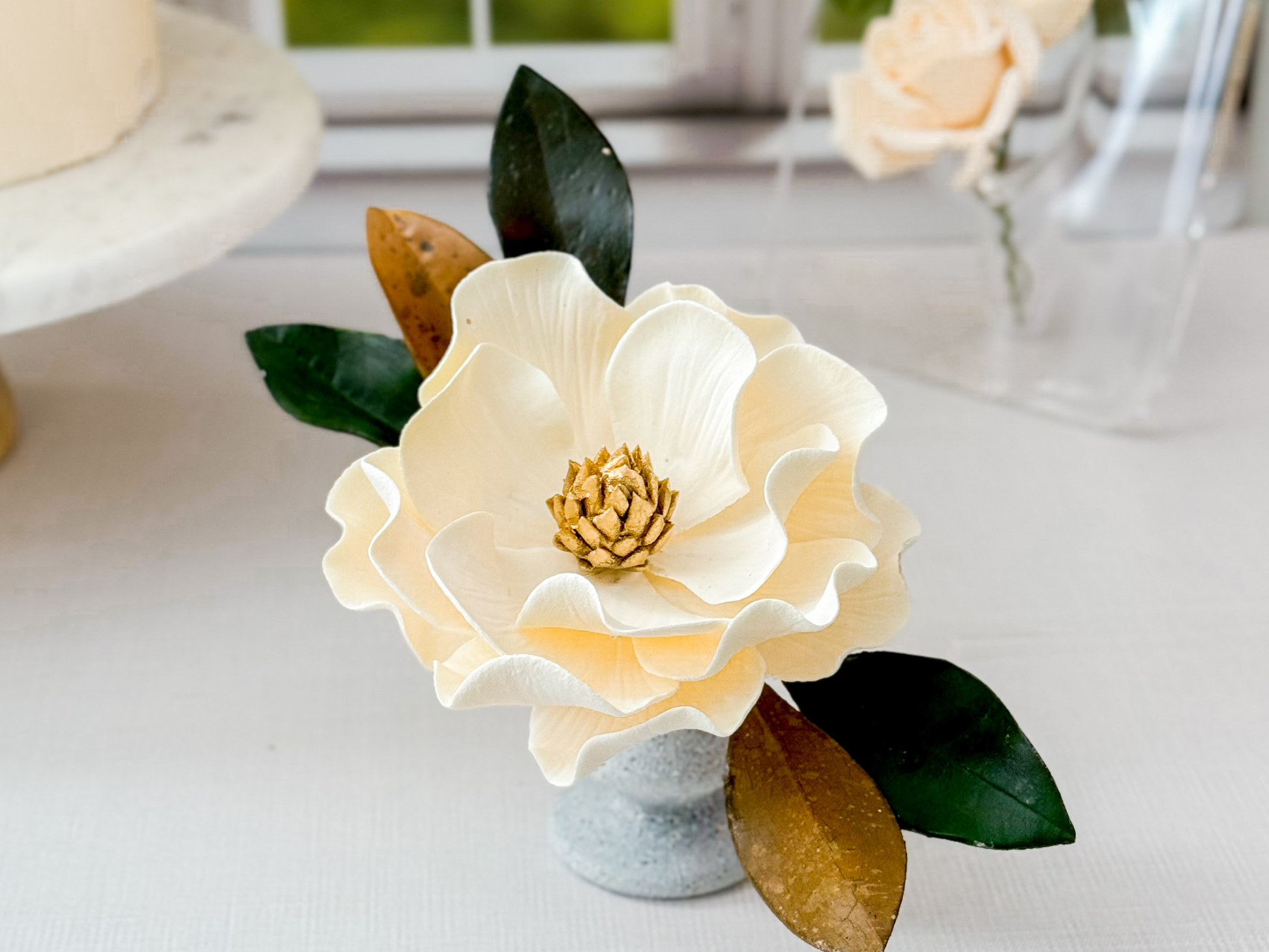 Decorative ivory sugar flower with a gold center and green and brown leaves on a white surface