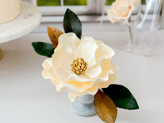 Decorative ivory sugar flower with a gold center and green and brown leaves on a white surface