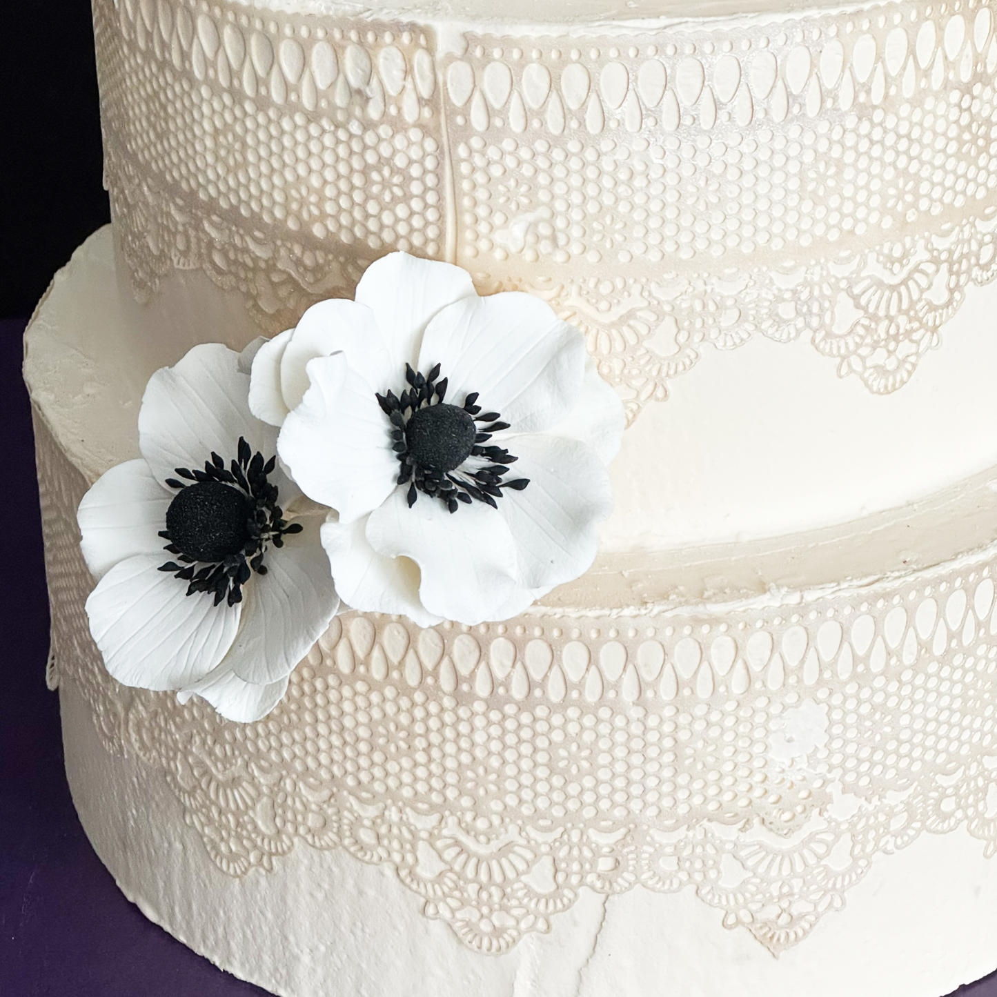 Close-up of a white lace wedding cake decorated using two sugar flower anemones with black centers on white petals.